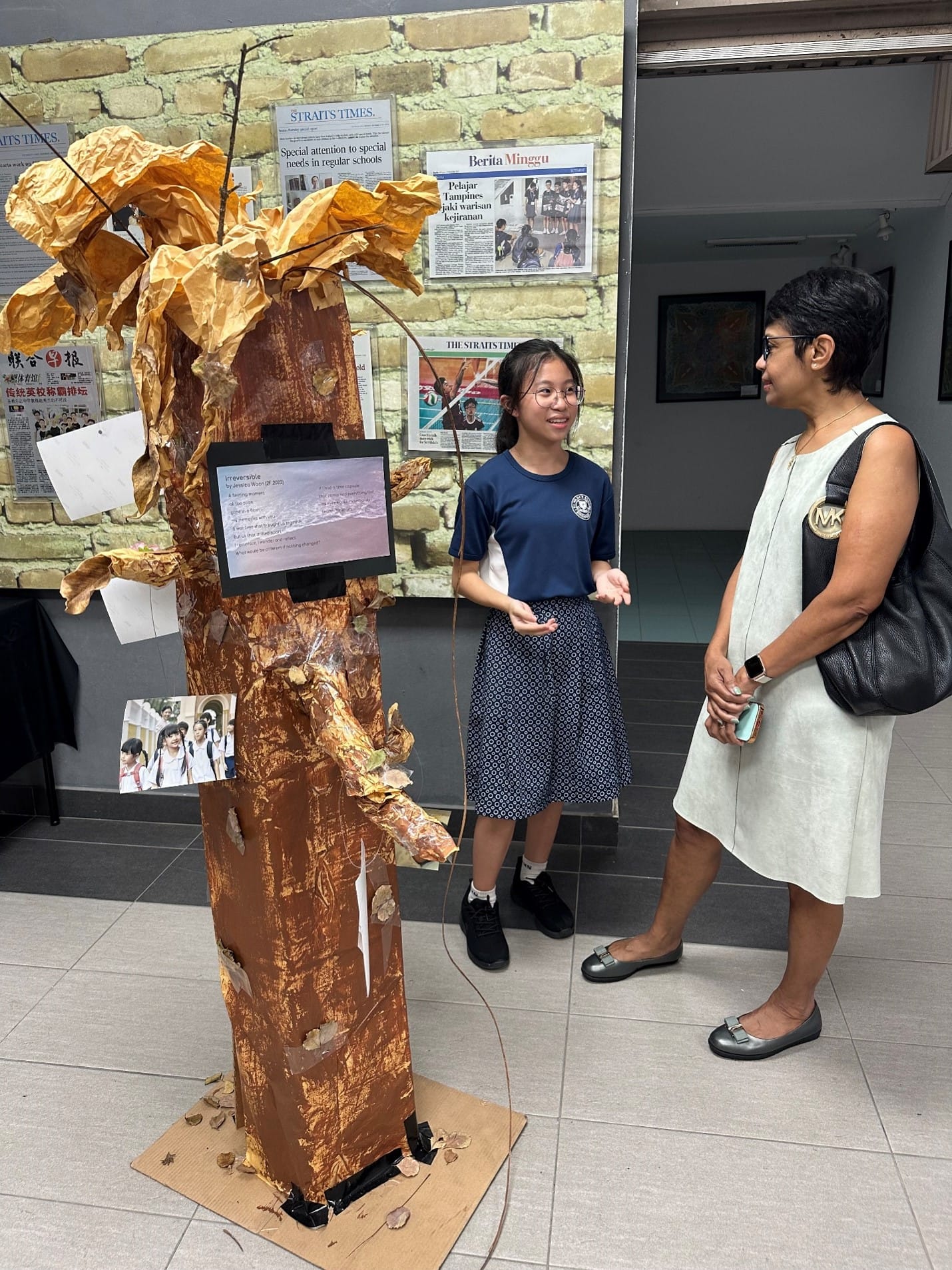 A young student in school uniform presents a handmade tree sculpture with a small screen to an adult woman at an exhibition.